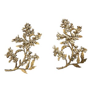 Pode incluir: Duas peças decorativas de arte de parede com um design de ramo floral. Os ramos são dourados com flores brancas e folhas verdes. As peças são feitas de metal e estão sobre um fundo branco.