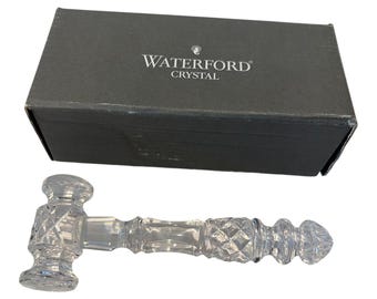 May include: A clear crystal gavel featuring intricate diamond-cut patterns on the head and handle, accompanied by a rectangular dark gray box with the white text WATERFORD CRYSTAL printed on the lid. The gavel has a polished, reflective surface.