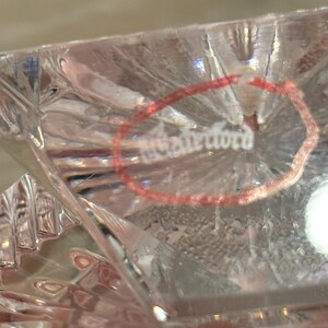 K&ouml;nnte beinhalten: Nahaufnahme eines klaren Kristallglases mit Diamantschliffmuster. Das Wort "Waterford" ist in einem roten Kreis sichtbar. Das Glas hat eine reflektierende Oberfl&auml;che.