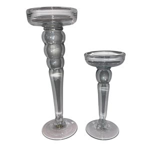May include: Two clear glass candle holders. The taller holder has a bulbous stem and a wide, flat top. The shorter holder has a similar design. Both are transparent and sit on a white background.