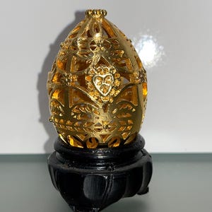 Puede incluir: Un huevo decorativo dorado con intrincados diseños de filigrana sobre un soporte de madera negro. El huevo es hueco y tiene un delicado patrón calado.
