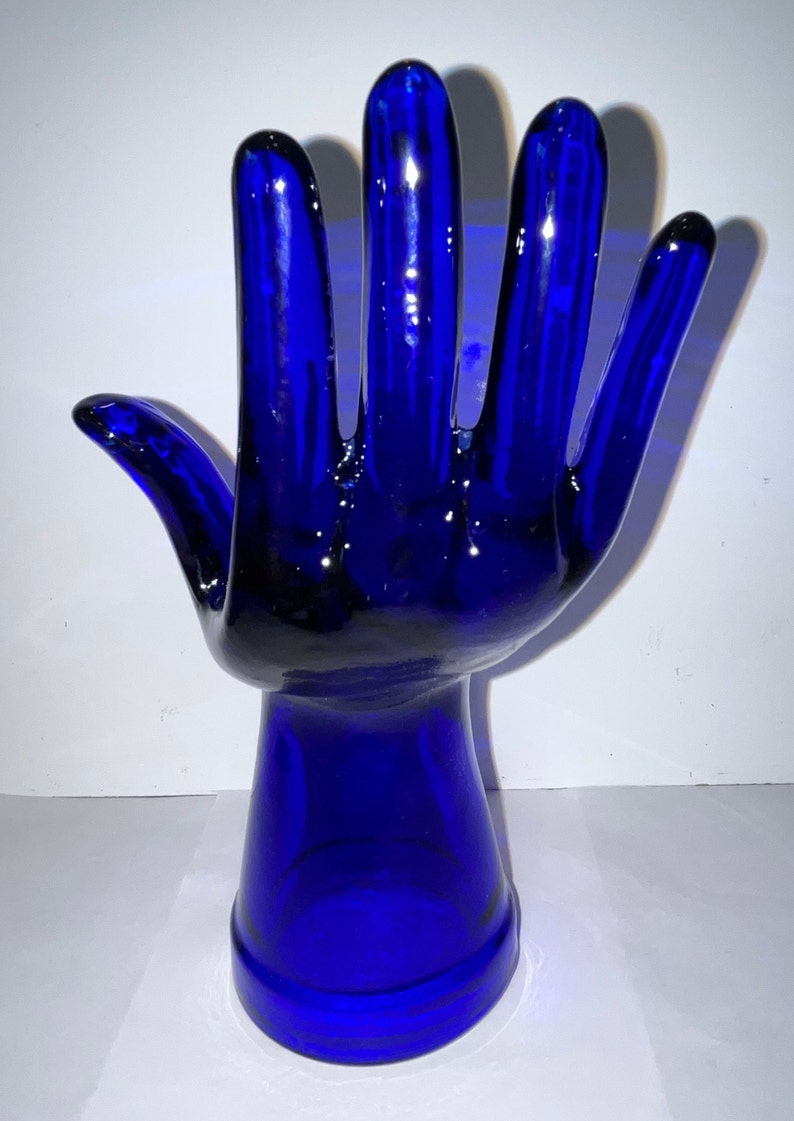 Peut inclure: Une sculpture de main en verre bleu cobalt avec une base arrondie. La main est lev&eacute;e, les doigts &eacute;cart&eacute;s.