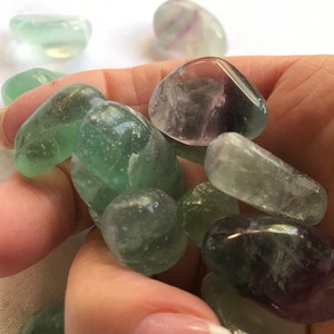 May include: A collection of tumbled green and purple fluorite stones. The stones are smooth and polished, with a variety of shades of green and purple.