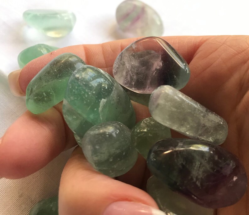 May include: A collection of tumbled green and purple fluorite stones. The stones are smooth and polished, with a variety of shades of green and purple.