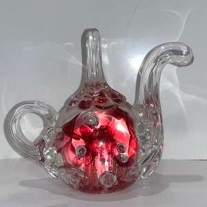 Puede incluir: Una tetera de vidrio transparente con un interior rojo lleno de burbujas. La tetera tiene un pico curvo y un asa.