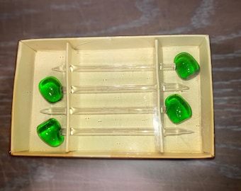 Pode incluir: Conjunto de quatro palitos de coquetel de vidro com pontas de vidro verde em uma caixa branca. Os palitos são transparentes e têm uma ligeira curva.