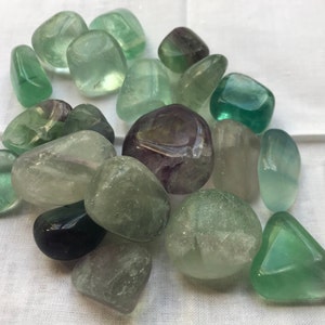 May include: A collection of tumbled fluorite stones in various shades of green, purple, and white. The stones are smooth and polished, with a translucent appearance.