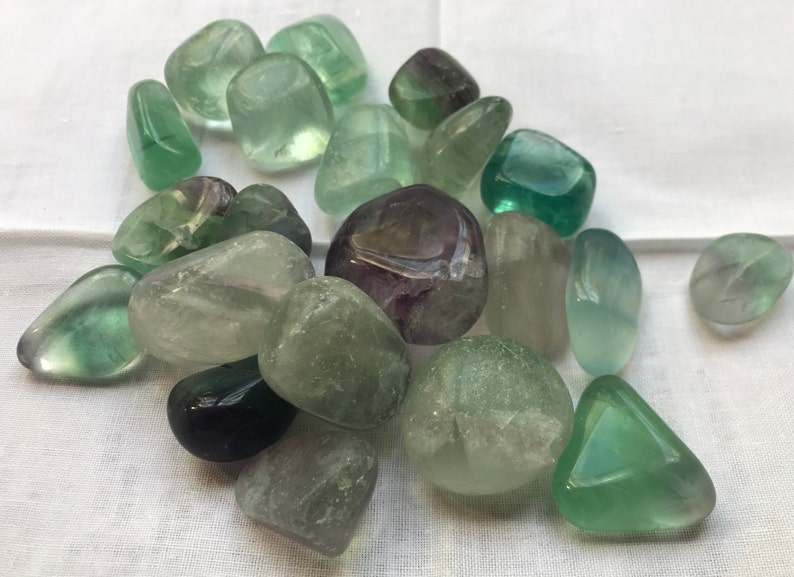 May include: A collection of tumbled fluorite stones in various shades of green, purple, and white. The stones are smooth and polished, with a translucent appearance.