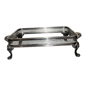Puede incluir: Una bandeja de servir rectangular de color plateado con un borde de celosía calado. La bandeja se apoya sobre cuatro patas curvas y ornamentadas con pies detallados, e incluye asas decorativas en los lados más cortos.
