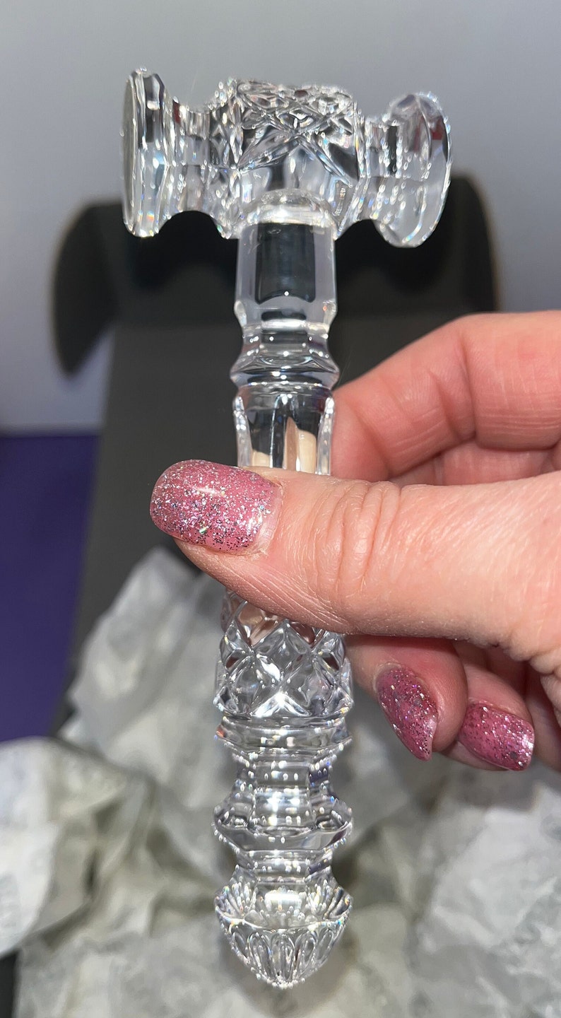 May include: A clear crystal gavel featuring intricate diamond-cut patterns on the handle and head. The head has a cylindrical shape with faceted ends, while the handle displays a multi-tiered, textured design with a pointed, decorative base.