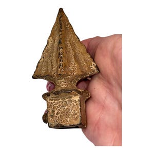 May include: A weathered, triangular-shaped metal finial with a pointed top and a square base. The object features a textured, distressed finish in shades of brown, tan, and cream, showing signs of oxidation and age across its entire surface.