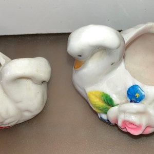 May include: Two white ceramic swan figurines with painted floral details. One swan has a pink interior and the other has a white interior.