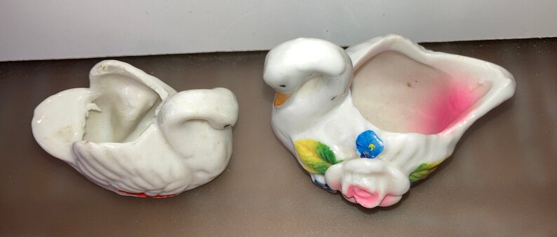 May include: Two white ceramic swan figurines with painted floral details. One swan has a pink interior and the other has a white interior.