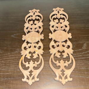 May include: Two unfinished wooden decorative appliques with intricate scrollwork and floral designs. Each piece features a central oval medallion and symmetrical patterns, perfect for woodworking projects or home decor.