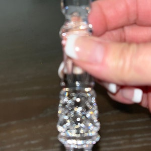 May include: A clear, crystal-like bottle stopper with a decorative, faceted design. The stopper has a cylindrical body and a flared top, all covered in sparkling, reflective facets. The overall effect is one of luxury and elegance.