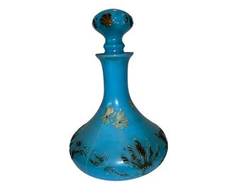 May include: A turquoise glass decanter with a matching stopper. The decanter features floral and leaf designs in cream and brown. The stopper has a seashell design in gold. The bottle is approximately 10 inches tall.