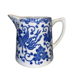 May include: White ceramic pitcher with a blue floral and bird pattern. The pitcher has a curved handle and a spout for pouring. The blue design includes flowers, swirls, and a stylized bird. The pitcher is likely for serving beverages.