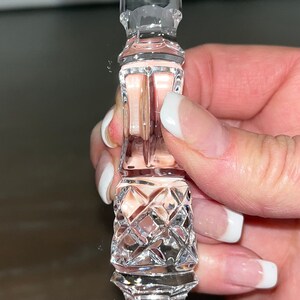 May include: A clear, faceted crystal bottle stopper with a decorative design. The stopper features a diamond-cut pattern and a cylindrical shape, perfect for wine bottles. The design is held by a hand.