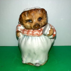 May include: A ceramic figurine of a hedgehog wearing a red and white plaid dress and carrying a small brown object. The figurine is white with brown accents and has a brown face with black eyes.