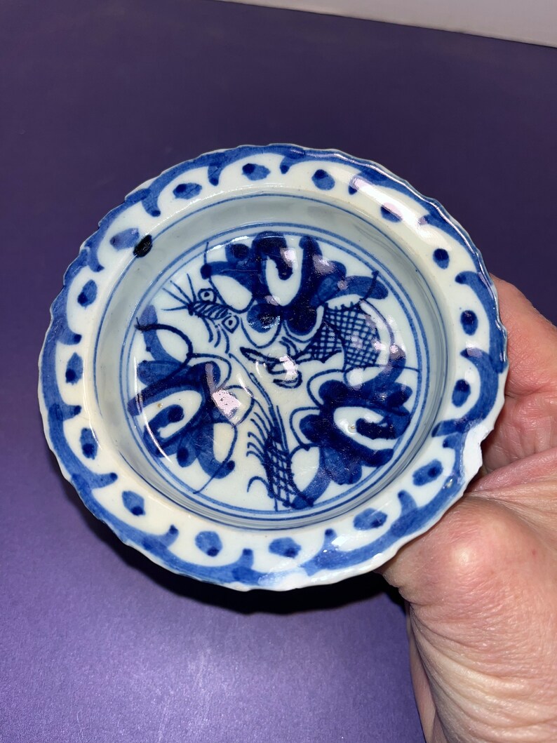 May include: A small, white ceramic bowl with a blue and white floral design. The bowl has a scalloped edge and a raised rim.