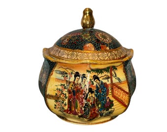 May include: A decorative ceramic jar with a lid. The jar features a hand-painted scene of figures in traditional attire, set against a landscape. The lid is adorned with floral patterns and a gold-coloured finial. The jar's body has a cream-coloured base.