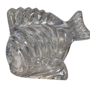 K&ouml;nnte beinhalten: Eine klare Fischskulptur aus Glas mit detaillierten, geschwungenen Linien. Der Fisch hat eine Flosse oben und eine Schwanzflosse. Die Skulptur ist transparent und reflektiert das Licht, wodurch ein schimmernder Effekt entsteht. Es ist ein dekorativer Gegenstand.