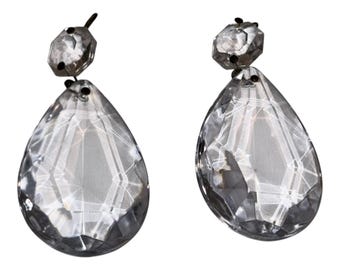 May include: A pair of clear, faceted crystal drop earrings. Each earring features a small, hexagonal top stone connected to a larger, teardrop-shaped pendant. The crystals have a reflective, multi-faceted surface that catches light.