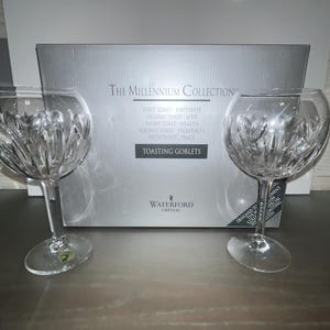 Op de afbeelding: Twee heldere kristallen Waterford toostglazen met een gesneden ontwerp. De glazen staan op een witte doos met de tekst "The Millennium Collection" en "Toasting Goblets".