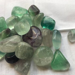 May include: A collection of green and purple fluorite tumbled stones. The stones are smooth and polished, with a variety of shades of green and purple.