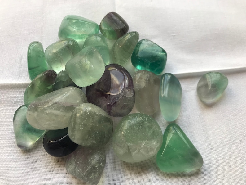 May include: A collection of green and purple fluorite tumbled stones. The stones are smooth and polished, with a variety of shades of green and purple.