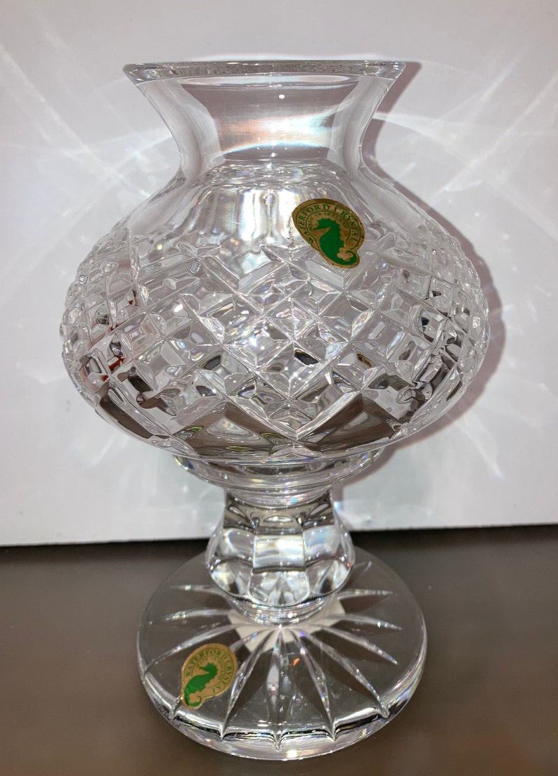 May include: A clear crystal vase with a cut glass design. The vase has a round body and a pedestal base. The vase has two labels on the bottom that say "Waterford Crystal" with a seahorse logo.