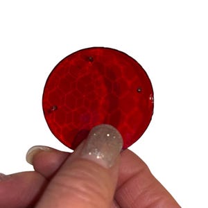 May include: A round, red object with a textured surface and three small holes. The object is held by a hand with a light-colored nail. The object has a honeycomb-like pattern.