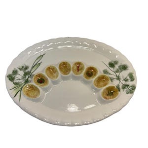 Puede incluir: Plato ovalado de cerámica blanca con borde festoneado. El plato presenta una sección central con siete huecos en forma de aceituna. Cada hueco contiene una aceituna pintada. Ramitas verdes de eneldo pintadas a ambos lados.