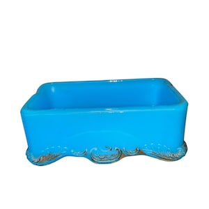 May include: A rectangular, turquoise-colored glass dish with a decorative, ornate base. The dish has a smooth, glossy finish and a simple, open design. The base features a gold-colored, scroll-like pattern.