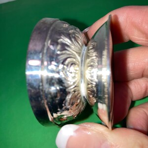 May include: A silver-colored, ornate, small dish with a decorative floral pattern. The dish has a round base and a flared, flat rim. The design is detailed and appears to be made of metal.