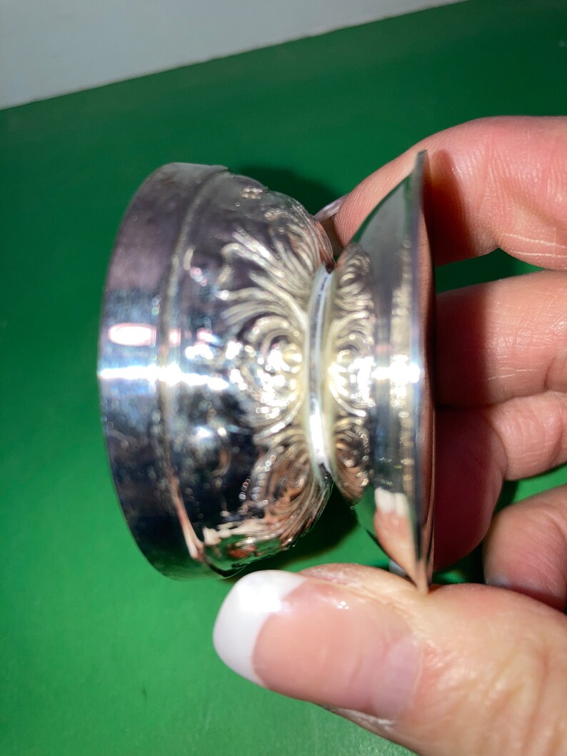 May include: A silver-colored, ornate, small dish with a decorative floral pattern. The dish has a round base and a flared, flat rim. The design is detailed and appears to be made of metal.