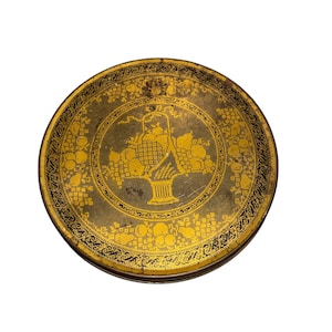 May include: Round, antique tin with a gold and black design. The lid features a central image of a basket overflowing with fruit, surrounded by a decorative border of flowers and vines. The tin has a slightly worn appearance.