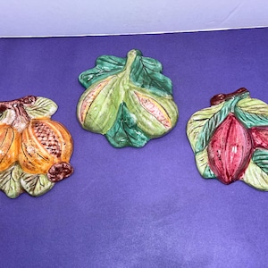 May include: Three ceramic wall plaques depicting fruit. The first plaque features two figs on a green leaf. The second plaque features two pomegranates on a green leaf. The third plaque features two red berries on a green leaf.