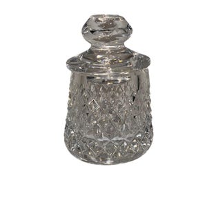 May include: A clear, cut-glass jar with a lid. The jar has a diamond pattern on the body and a rounded base. The lid has a knob on top. The jar is likely used for storage or display.