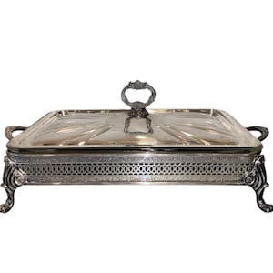 Puede incluir: Una fuente de servir rectangular de metal plateado con tapa a juego. La fuente presenta patas curvas ornamentadas, un borde decorativo de celosía calada y dos asas laterales. La tapa tiene un asa central con detalles florales.