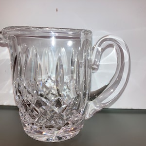May include: A clear glass creamer with a cut crystal design. The creamer has a handle and a rounded base.