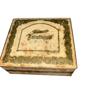May include: Vintage rectangular tin box with a cream-colored lid and sides. The lid features the words "Whitman's" and "Loneliness" in an ornate font, framed by green and gold decorative borders. The box has a distressed, aged appearance.