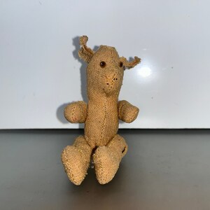 Op de afbeelding: Een klein, bruin, handgemaakt pluche speelgoed in de vorm van een beer. De beer heeft een gestikte neus en kleine, bruine ogen.
