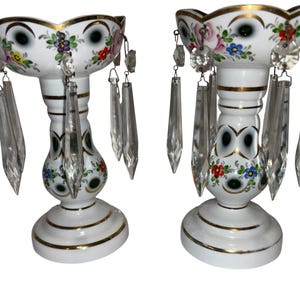 Może przedstawiać: Para białych porcelanowych świeczników z kwiatowymi wzorami i złotymi obramowaniami. Każdy świecznik ma wiszące kryształowe pryzmaty. Świeczniki mają falisty brzeg i podstawę.