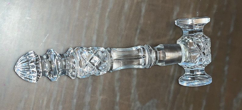 May include: A clear, cut-glass gavel with a decorative handle and head. The handle features diamond and geometric patterns, and the head has a rectangular shape. The gavel is designed for ceremonial use.