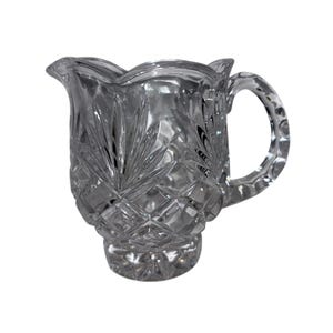 May include: A clear crystal creamer with a decorative handle and a flared, scalloped rim. The creamer features a diamond and leaf pattern, adding a touch of elegance to any table setting. The creamer is approximately 5 inches tall.