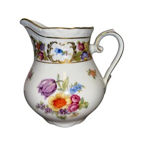 May include: A white porcelain pitcher with a bulbous body and a curved handle. The pitcher is decorated with colorful floral designs, including a large purple tulip, and gold trim. The rim has a scalloped edge.