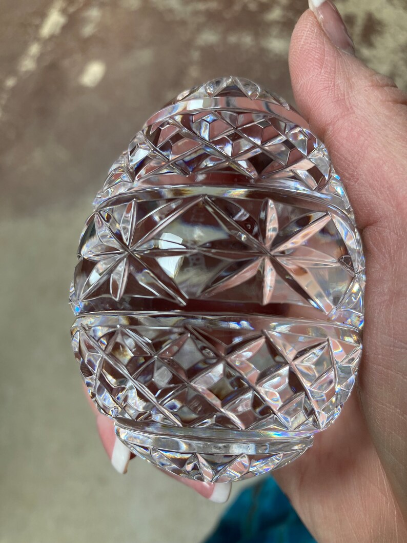 K&ouml;nnte beinhalten: Ein klarer, eif&ouml;rmiger Dekorationsgegenstand aus geschliffenem Glas. Das Ei weist ein detailliertes geometrisches Muster aus Rauten und Sternen auf. Die Facetten fangen das Licht ein und erzeugen einen funkelnden Effekt. Es ist wahrscheinlich ein Dekorationsartikel.