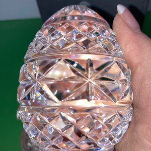 K&ouml;nnte beinhalten: Ein klarer, facettierter Kristallei mit einem Diamanten- und Sternmuster. Das Ei wird in einer Hand gehalten, wodurch die komplizierten Schnitte, die das Licht reflektieren, zur Geltung kommen. Das dekorative Objekt ist ein sch&ouml;nes St&uuml;ck Wohnkultur.