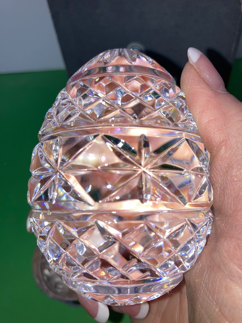 K&ouml;nnte beinhalten: Ein klarer, facettierter Kristallei mit einem Diamanten- und Sternmuster. Das Ei wird in einer Hand gehalten, wodurch die komplizierten Schnitte, die das Licht reflektieren, zur Geltung kommen. Das dekorative Objekt ist ein sch&ouml;nes St&uuml;ck Wohnkultur.